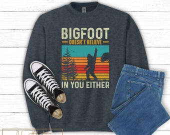 May include: Dark gray sweatshirt with the text "BIGFOOT DOESN'T BELIEVE IN YOU EITHER" above a graphic of Bigfoot in a sunset scene. The image includes black and white sneakers and blue jeans.
