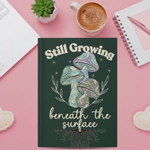 May include: A dark green hardcover journal featuring a central illustration of three textured mushrooms in shades of blue, purple, and cream. The text Still Growing is printed above the mushrooms, and beneath the surface is printed below.