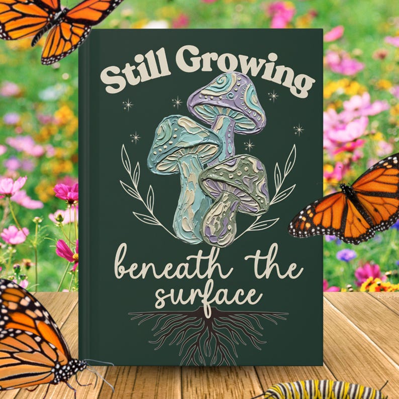 May include: A dark green hardcover book featuring a textured illustration of three mushrooms in shades of blue, purple, and green. The text "Still Growing" appears above the mushrooms, and "beneath the surface" is printed below with root graphics.