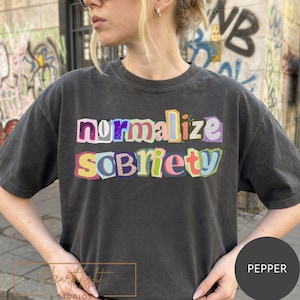 May include: A dark gray t-shirt with the words "normalize sobriety" in colorful, cut-out style letters. The shirt is a casual style, with a crew neck and short sleeves. The shirt is a pepper color.