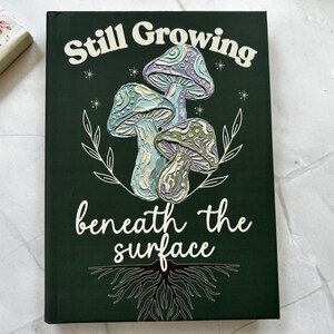 May include: A dark green hardcover book featuring a textured illustration of three mushrooms in shades of purple, blue, and green. The text Still Growing is printed above the mushrooms, and beneath the surface is printed below with root graphics.