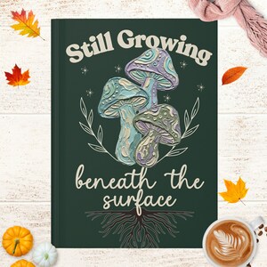 May include: A dark green rectangular journal cover featuring a central illustration of three textured mushrooms in shades of blue, purple, and green. The text "Still Growing" and "beneath the surface" is printed in cream, with root illustrations below.