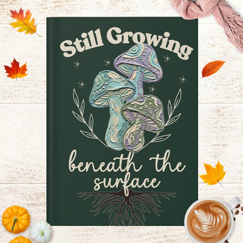 May include: A dark green rectangular journal cover featuring a central illustration of three textured mushrooms in shades of blue, purple, and green. The text "Still Growing" and "beneath the surface" is printed in cream, with root illustrations below.