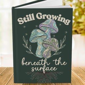 May include: A dark green hardcover journal featuring a central illustration of three textured mushrooms in shades of blue, purple, and cream. The text Still Growing is printed above the mushrooms, and beneath the surface is printed below.