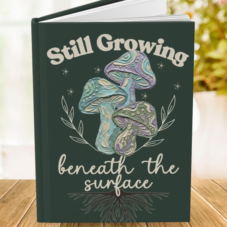 May include: A dark green hardcover journal featuring a central illustration of three textured mushrooms in shades of blue, purple, and cream. The text Still Growing is printed above the mushrooms, and beneath the surface is printed below.