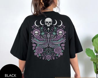 May include: Black t-shirt featuring a detailed moth and skull design. The graphic includes a moth with teal and purple wings, skulls, crescent moons, and floral elements. The word "BLACK" is printed on the lower left.