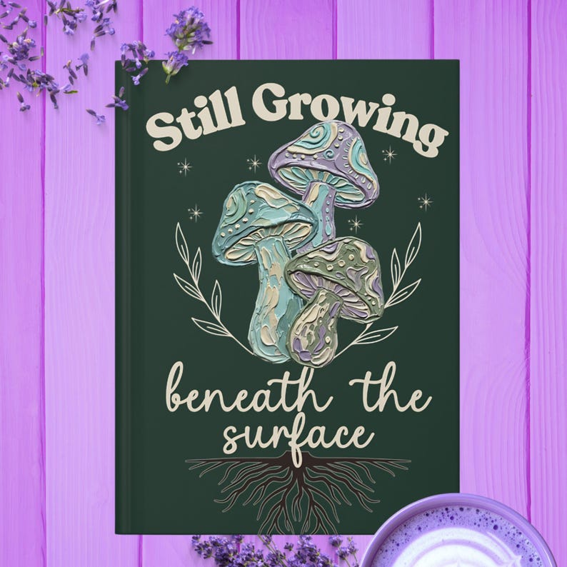 May include: A dark green notebook featuring a central illustration of three textured mushrooms in shades of blue, purple, and cream. The text "Still Growing" appears above and "beneath the surface" below, accompanied by white line art of leaves and roots.