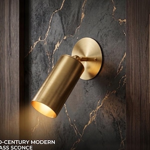 Può includere: Applique in ottone moderno di metà secolo con un apparecchio cilindrico e un supporto a parete circolare. L'applique è montata su una parete grigio scuro con venature dorate. Il testo "MID-CENTURY MODERN BRASS SCONCE" è in basso.