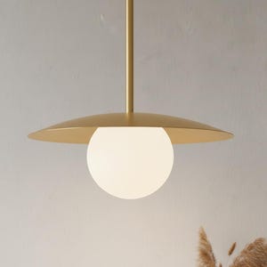 May include: A modern pendant light fixture with a gold finish. The fixture features a circular, flat shade above a spherical, white glass globe. The light hangs from a slender, gold-colored rod.
