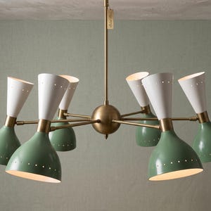 May include: A multi-arm chandelier featuring six cone-shaped shades with a two-tone finish of sage green and white. The fixture has a central brass-toned sphere and matching arms, with small perforated holes along the rim of each metal shade.