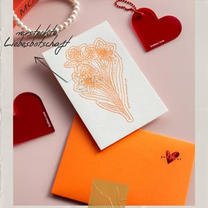 Könnte beinhalten: Eine Grußkarte mit einem orangefarbenen Umschlag, einer weißen Karte mit einem orangefarbenen Blumendesign und zwei roten herzförmigen Acrylanhängern. Der Text auf der Karte lautet "rersteckte Liebesbotschaft". Das Bild befindet sich auf einem rosa Hintergrund.