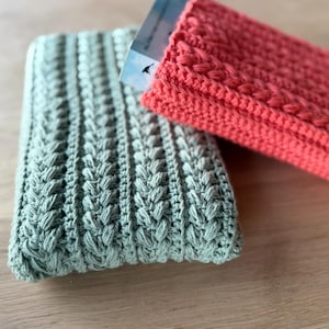 May include: Two handmade crochet cases in soft colors. One is a muted green, the other a coral pink. The cases have a textured, raised pattern. One case holds a partially visible card or paper. The cases are likely designed to hold small items.