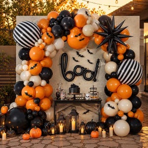 May include: A Halloween balloon arch featuring orange, black, and cream balloons, a large black starburst balloon, and black striped spheres. The display includes a black cursive 'boo' balloon, bat cutouts, and a table with lanterns and pumpkins.