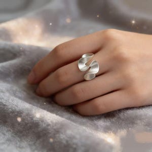 May include: A silver-colored ring with a unique, flowing design. The ring features two curved, ribbon-like elements that overlap, creating an abstract, modern look. The ring is worn on a finger, showcasing its elegant form.