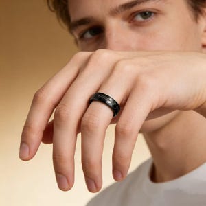 以下が含まれることがあります： 彫刻されたテキストとデザインが施された黒い指輪。指にはめられています。リングは濃い色で、金属製のように見えます。リングはバンドスタイルです。