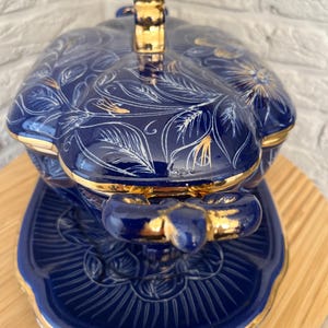 May include: A blue ceramic serving dish with a lid, featuring gold accents and white floral and leaf patterns. The dish sits on a matching saucer, creating a cohesive set. The handle and edges are trimmed in gold.