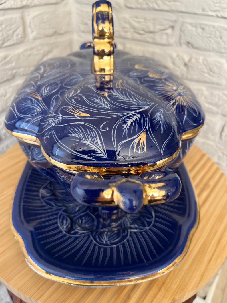 May include: A blue ceramic serving dish with a lid, featuring gold accents and white floral and leaf patterns. The dish sits on a matching saucer, creating a cohesive set. The handle and edges are trimmed in gold.
