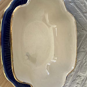 May include: Cream-coloured, scalloped-edge serving dish with gold trim and handles, on a navy blue plate with gold accents. The dish has a glossy finish, suggesting ceramic or porcelain material.
