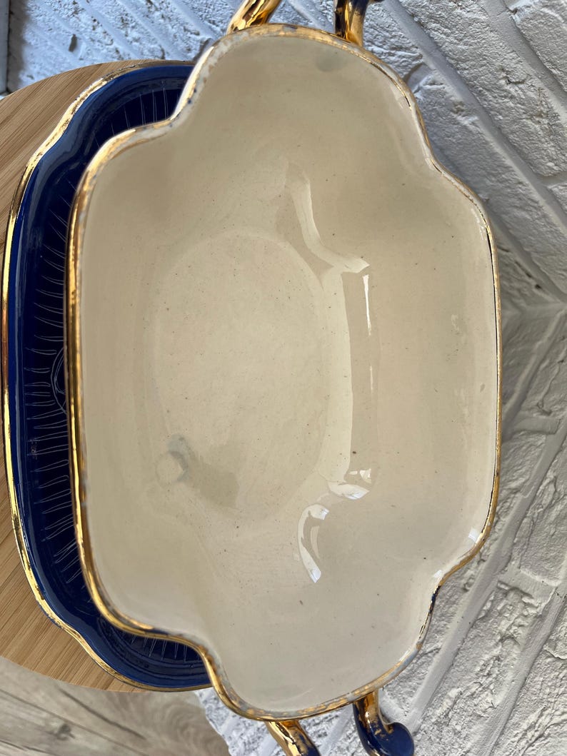 May include: Cream-coloured, scalloped-edge serving dish with gold trim and handles, on a navy blue plate with gold accents. The dish has a glossy finish, suggesting ceramic or porcelain material.
