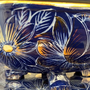 May include: A dark blue ceramic bowl with a gold rim and three feet. The bowl is decorated with white and gold floral designs. The bowl is sitting on a clear glass surface.