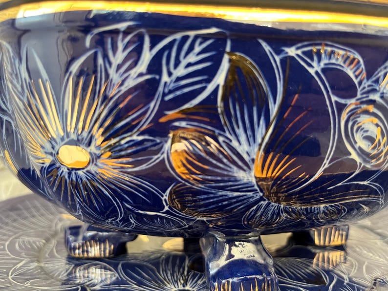 May include: A dark blue ceramic bowl with a gold rim and three feet. The bowl is decorated with white and gold floral designs. The bowl is sitting on a clear glass surface.