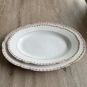 Op de afbeelding: Twee witte ovale serveerschalen met een rood en gouden decoratieve rand. De schalen zijn op elkaar gestapeld. De schalen hebben een geschulpte rand. Ideaal voor het serveren van voedsel.
