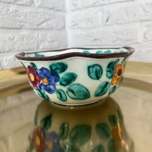Könnte beinhalten: Eine kleine, dekorative Keramikschale mit gewelltem Rand und floralem Design. Die Schale hat eine cremefarbene Basis mit handgemalten Blumen in Rot-, Blau- und Orangetönen sowie grünen Blättern. Der Rand ist dunkelbraun.