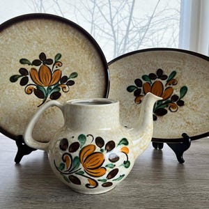 May include: A cream-coloured ceramic teapot and two serving platters with a floral design. The teapot and platters have brown rims and feature orange, green, and brown floral patterns. The teapot has a handle and spout.