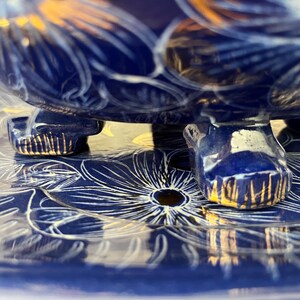 May include: Close-up of a blue ceramic bowl with gold accents and floral designs. The bowl rests on a matching plate, also decorated with white floral patterns. The bowl's base has three feet with gold trim.