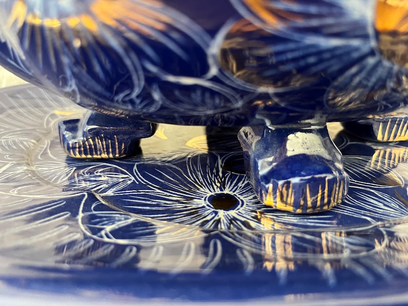 May include: Close-up of a blue ceramic bowl with gold accents and floral designs. The bowl rests on a matching plate, also decorated with white floral patterns. The bowl's base has three feet with gold trim.