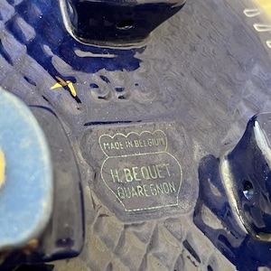 May include: Close-up of a deep blue ceramic object with a textured surface. The base features a rectangular stamp that reads "MADE IN BELGIUM" and "H. BEQUET QUAREGNON". The object has a light blue accent.