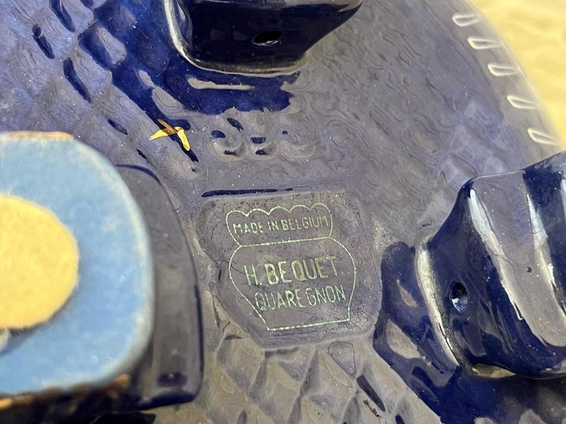 May include: Close-up of a deep blue ceramic object with a textured surface. The base features a rectangular stamp that reads "MADE IN BELGIUM" and "H. BEQUET QUAREGNON". The object has a light blue accent.
