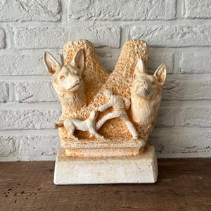 Può includere: Scultura beige raffigurante due teste di cane, un cagnolino e una persona. La scultura è posta su una base rettangolare. Lo sfondo è un muro di mattoni bianchi.