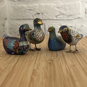 Può includere: Una collezione di quattro piccole statuette di uccelli realizzate con un intricato lavoro di smalto cloisonné. Ogni pezzo presenta vivaci motivi multicolori, dettagli in filo metallico dorato e distinte forme di uccelli.