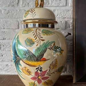 Può includere: Un vaso in ceramica color crema con coperchio, con un disegno di uccello dipinto a mano con accenti verdi, gialli e rossi. Il vaso ha finiture dorate e un manico dorato sul coperchio. L'uccello è circondato da motivi floreali e fogliari.