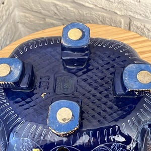 May include: Underside view of a dark blue ceramic bowl with white floral patterns. The bowl has four square feet with gold pads. The base features a textured design and the text "BEDUCT".