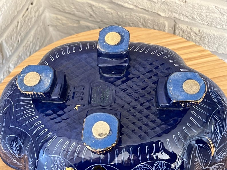 May include: Underside view of a dark blue ceramic bowl with white floral patterns. The bowl has four square feet with gold pads. The base features a textured design and the text "BEDUCT".