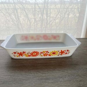 Puede incluir: Fuente rectangular blanca para horno con un diseño floral en naranja, amarillo y rojo. La fuente tiene un interior y asas de color gris claro. El patrón floral se repite a lo largo de los bordes superior e inferior.