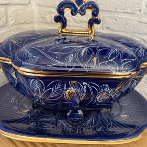 May include: A navy blue ceramic serving dish with a lid, featuring gold trim and handles. The dish is decorated with white floral and leaf patterns. It sits on a matching plate, creating a cohesive set.