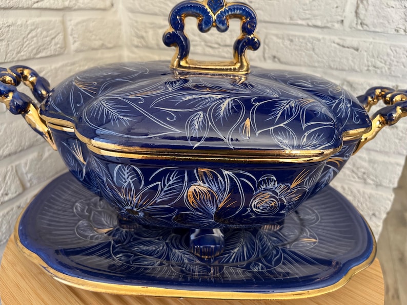 May include: A navy blue ceramic serving dish with a lid, featuring gold trim and handles. The dish is decorated with white floral and leaf patterns. It sits on a matching plate, creating a cohesive set.