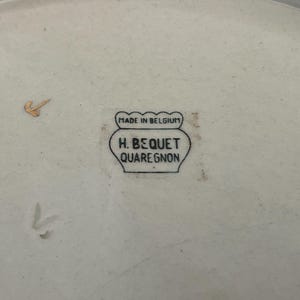 May include: A close-up of a cream-coloured ceramic plate with a gold accent. The plate features a black stamp that reads "MADE IN BELGIUM" above "H. BEQUET QUAREGNON". The plate has a slightly irregular shape.
