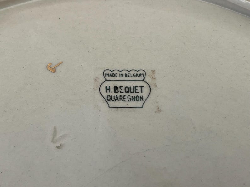 May include: A close-up of a cream-coloured ceramic plate with a gold accent. The plate features a black stamp that reads "MADE IN BELGIUM" above "H. BEQUET QUAREGNON". The plate has a slightly irregular shape.
