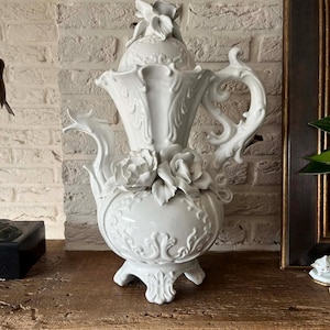 Op de afbeelding: Een witte keramische theepot met ingewikkelde bloemen- en krulwerkdetails. De theepot heeft een grote handgreep, een decoratieve tuit en een deksel met een bloem. De basis heeft drie sierlijke voetjes. Het is een decoratief item.