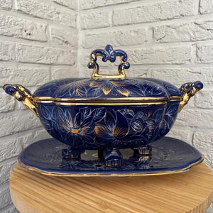 May include: A navy blue ceramic serving dish with a lid, decorated with gold floral patterns and accents. The dish rests on a matching plate with a decorative base. Gold handles and lid finial complete the design.