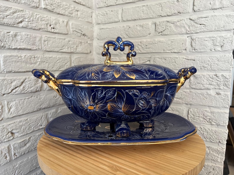May include: A navy blue ceramic serving dish with a lid, decorated with gold floral patterns and accents. The dish rests on a matching plate with a decorative base. Gold handles and lid finial complete the design.