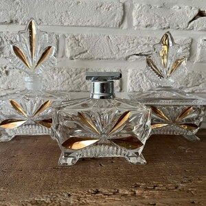 May include: Three vintage glass perfume bottles with gold accents. The bottles have a clear, faceted design and are topped with decorative stoppers. The gold leaf-like accents add a touch of elegance to the set.