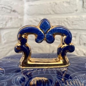 May include: Close-up of a blue and gold ceramic lid with a decorative handle. The handle is a deep blue with gold accents. The lid has a floral pattern in white and gold.