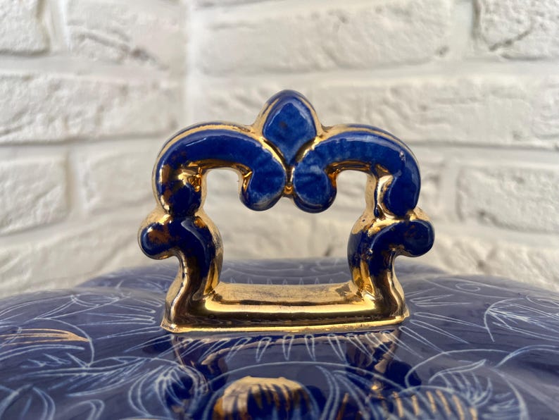 May include: Close-up of a blue and gold ceramic lid with a decorative handle. The handle is a deep blue with gold accents. The lid has a floral pattern in white and gold.