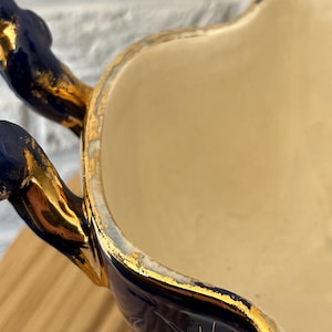 May include: Close-up of a vintage ceramic serving dish. The dish has a cream-coloured interior and a navy blue exterior with gold accents. The handles are navy blue with gold trim, and the rim is also trimmed in gold.