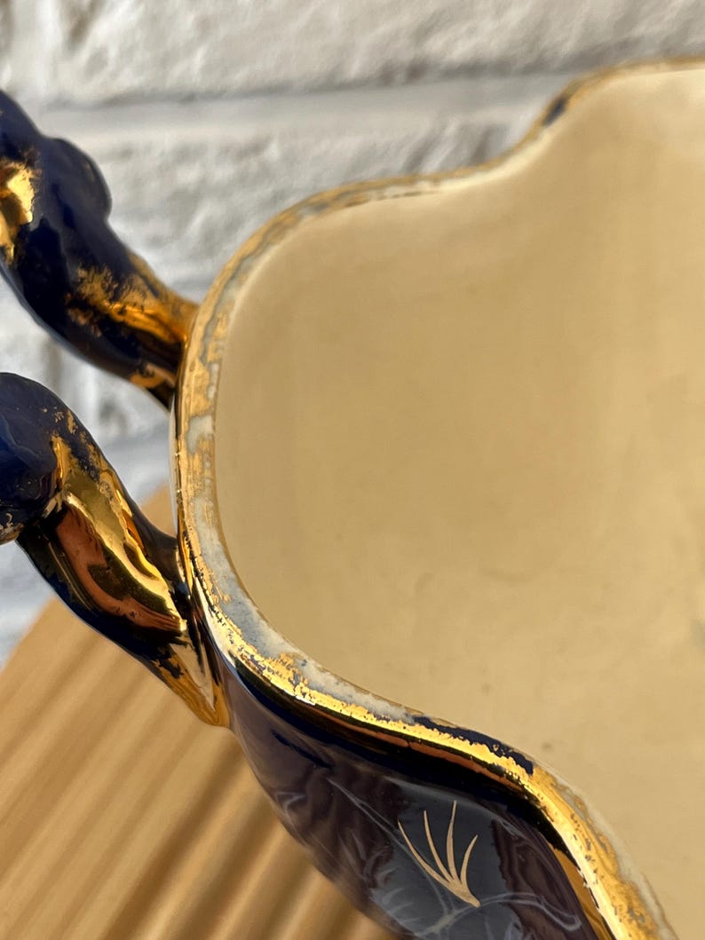 May include: Close-up of a vintage ceramic serving dish. The dish has a cream-coloured interior and a navy blue exterior with gold accents. The handles are navy blue with gold trim, and the rim is also trimmed in gold.
